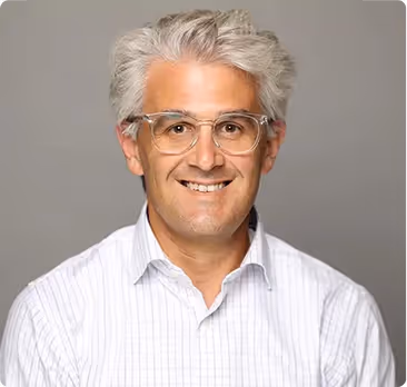 Smiling middle-aged man with gray hair and clear glasses wearing a white striped shirt against a gray background.