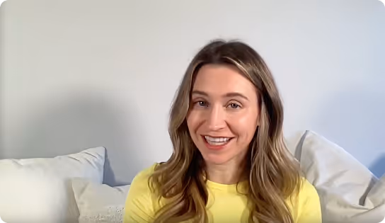 Smiling woman with long wavy hair wearing a yellow top sitting on a white sofa with cushions.