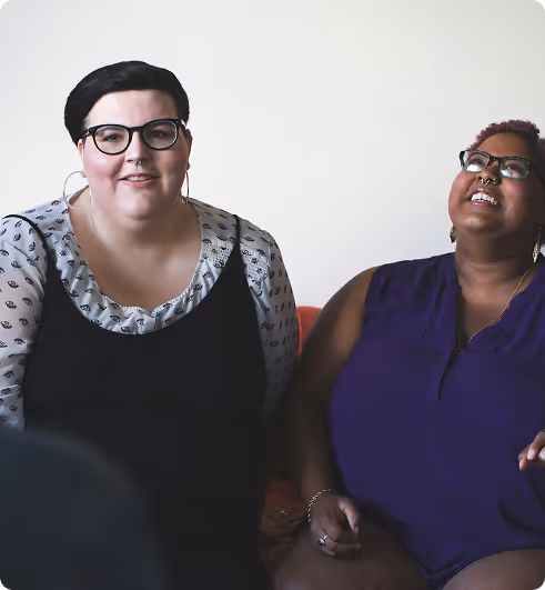 Two women sitting together, one with glasses and large hoop earrings smiling, the other wearing glasses and a purple top laughing while looking up.