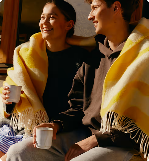 A smiling young couple sitting close, wrapped in a yellow and white striped blanket, each holding a white cup.