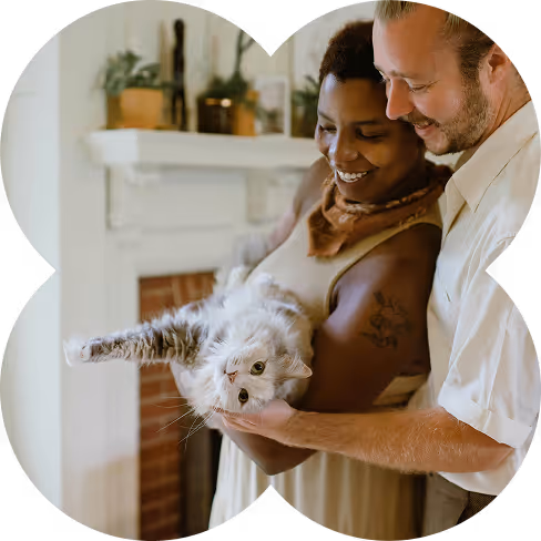 Smiling couple holding and petting a relaxed white and gray cat indoors near a fireplace.