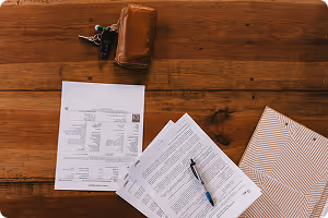 Wooden table with documents, a pen, a wallet, and a set of keys placed on it.