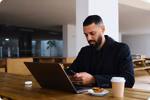 Man in black jacket sitting at a wooden table using a smartphone with a laptop, coffee cup, and pastry nearby.