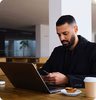 Man with beard and dark hair sitting at a table using a smartphone with a laptop open in front of him and a coffee cup nearby.