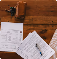 Wooden table with a leather wallet and keys, printed documents, and a blue pen on top of paper.