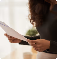 Person with curly hair holding and reviewing papers indoors near window light.