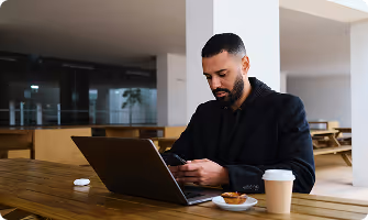 Man in black jacket using smartphone with laptop and coffee on wooden table in outdoor seating area.