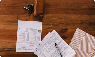 Wooden table with house keys, wallet, printed documents, pen, and a patterned folder.
