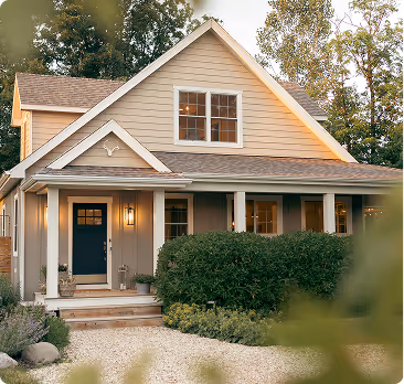 Cozy beige two-story house with a blue front door, lit porch lights, and surrounding greenery.