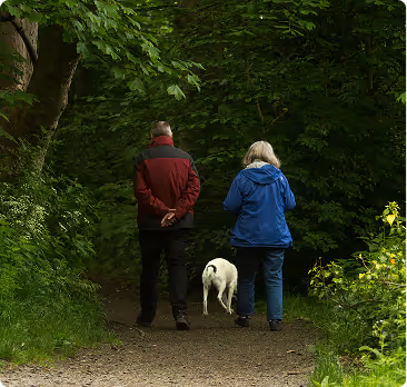 Two people walking on a forest path with a white dog, surrounded by dense green foliage.