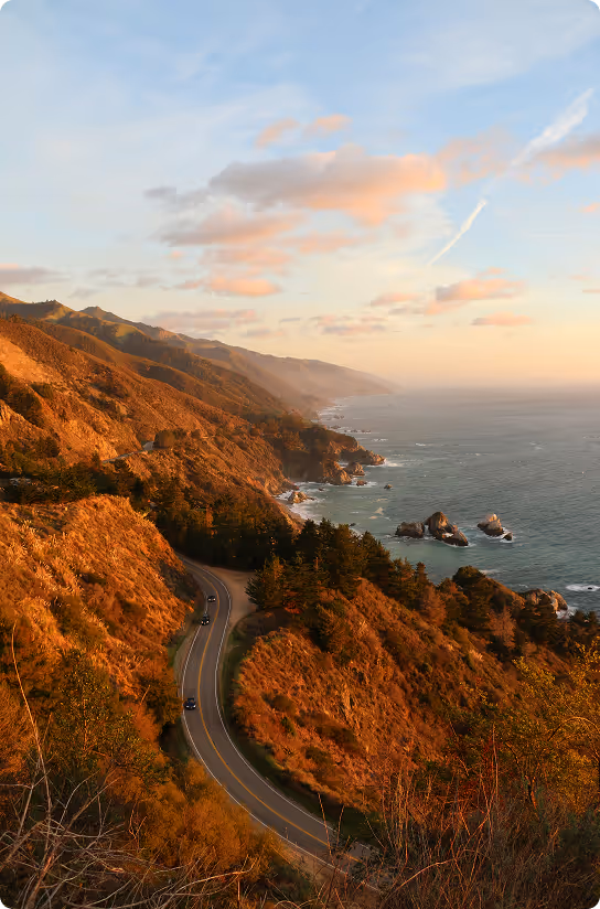 Coastal highway winding through sunlit mountains with ocean and rocky shore under a partly cloudy sky at sunset.