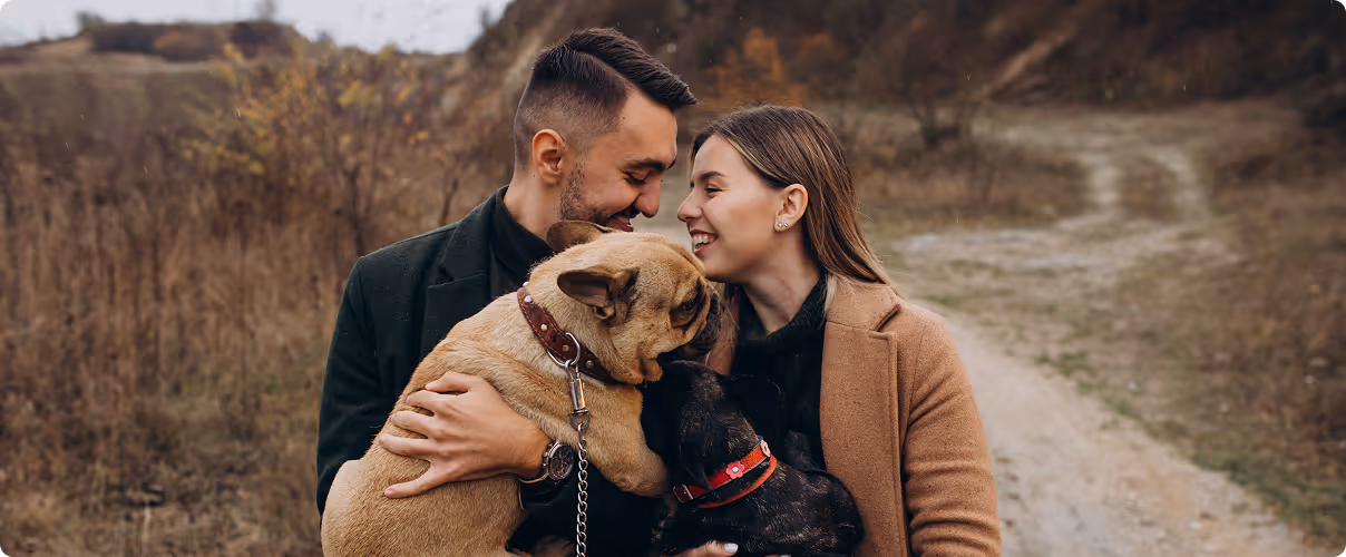 Smiling couple outdoors embracing two French bulldogs on a dirt path in a natural setting.