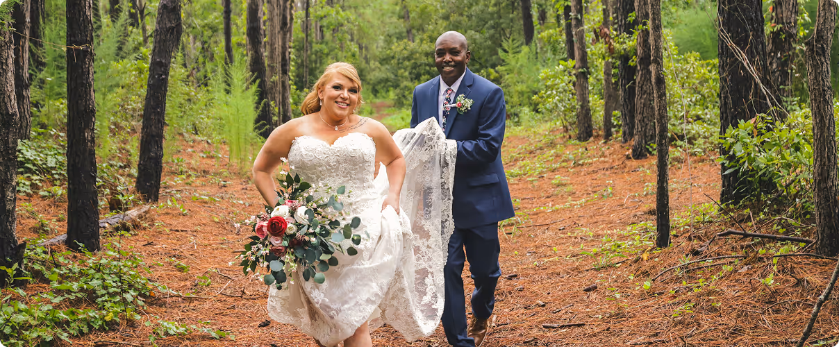 Bride holding bouquet and lifting wedding dress running ahead of groom in a forest pathway.
