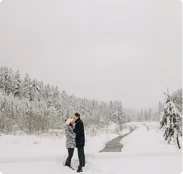 Couple embracing and kissing in a snowy landscape with snow-covered trees and a frozen river.