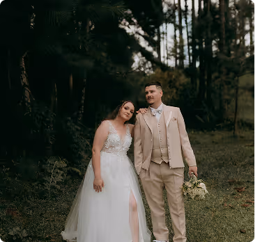 Bride in a white sleeveless gown leaning on groom in a beige suit holding a bouquet in a forest setting.
