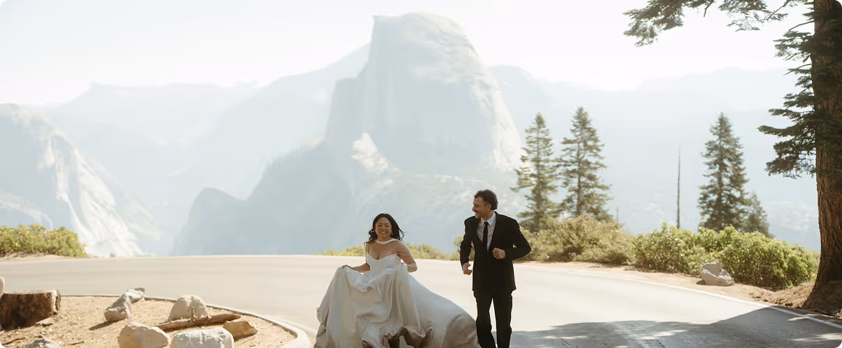 Bride in a white dress and groom in a black suit running joyfully on a mountain road with trees and rocky cliffs in the background.