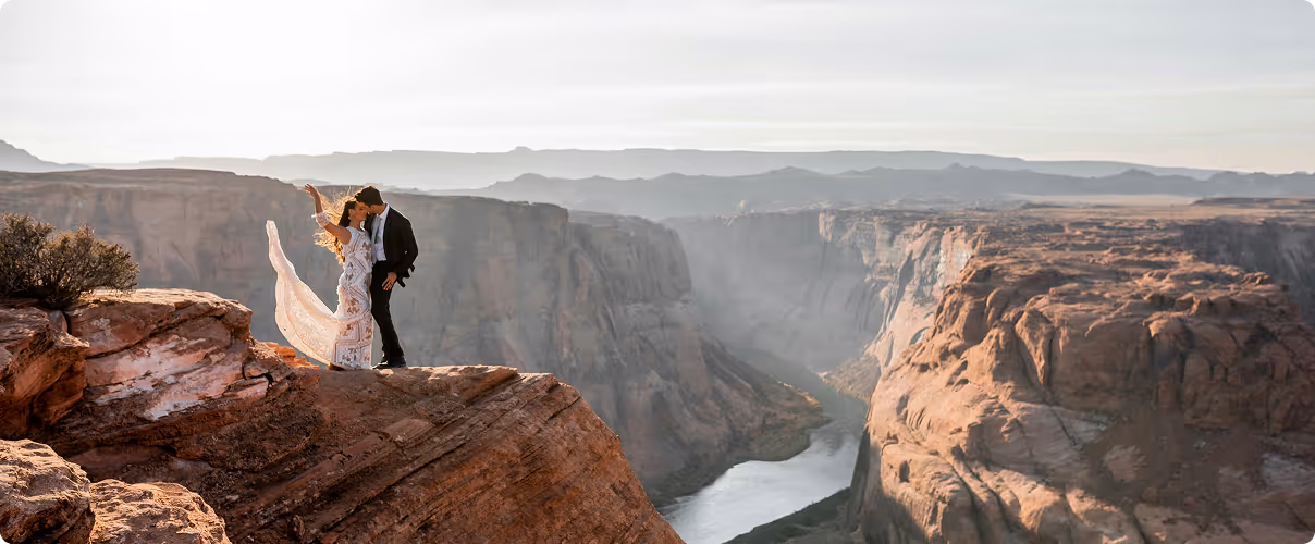Couple in formal attire standing on a cliff edge overlooking a river winding through a canyon at sunset.