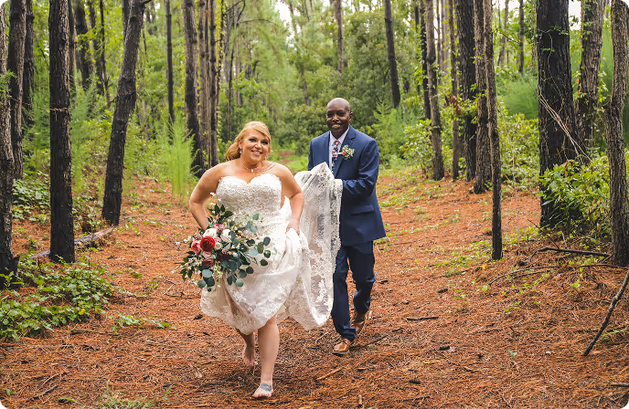 Bride in a white wedding dress holding a bouquet running ahead of the groom in a navy suit on a forest path.