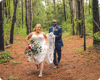 Smiling bride in white wedding dress holding bouquet running with groom in navy suit through a forest path.