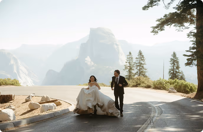 Bride and groom joyfully running on a mountain road with scenic cliffs and trees in the background.