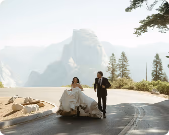 Bride and groom walking hand in hand on a mountain road with scenic cliffs and trees in the background.