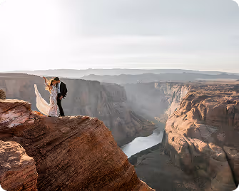 Bride and groom standing on the edge of a rocky cliff overlooking a winding river in a deep canyon at sunset.