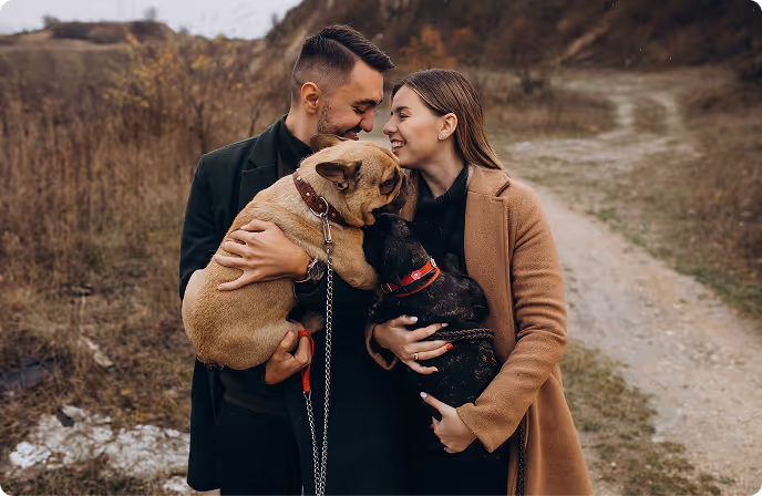 Smiling couple outdoors on a dirt path, each holding a dog and leaning their heads close together.