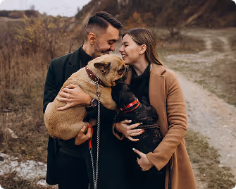 Couple outdoors on a dirt path, each holding a dog and smiling at each other.