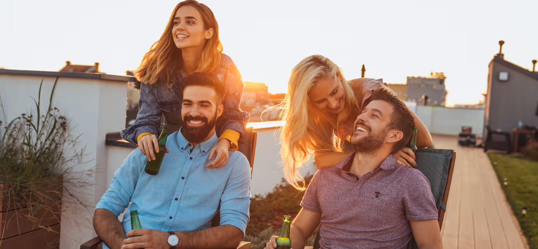 Two couples enjoying drinks on a rooftop at sunset, with women standing behind seated men, all smiling and relaxed.