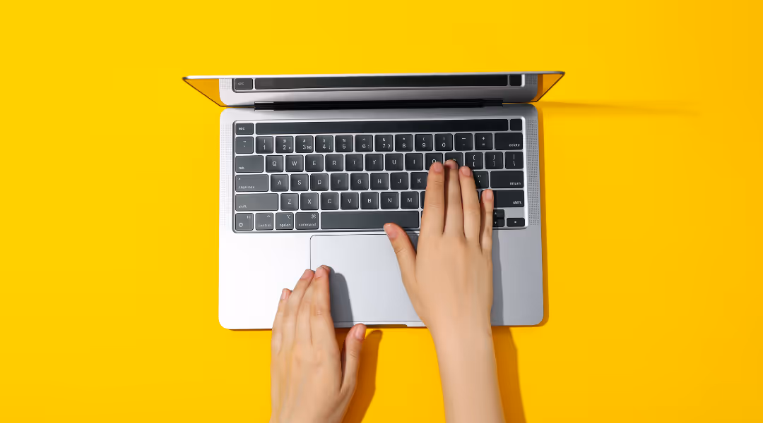 Hands typing on a laptop keyboard with a bright yellow background.