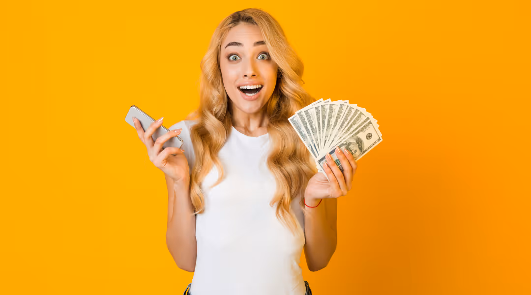Excited young woman holding a smartphone in one hand and fanned out US dollar bills in the other against an orange background.