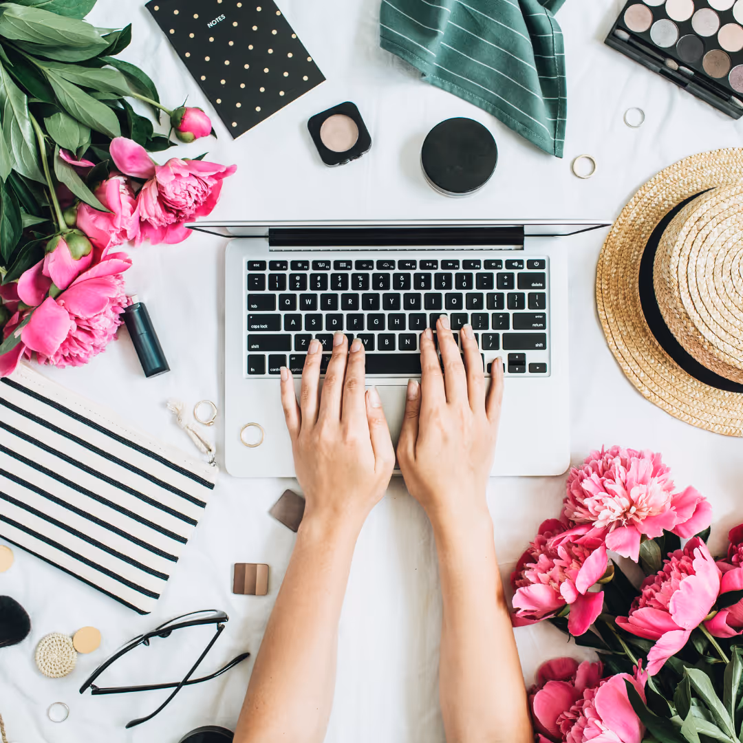 Hands typing on a laptop keyboard surrounded by pink flowers, makeup items, glasses, a striped pouch, a hat, and a green cloth.