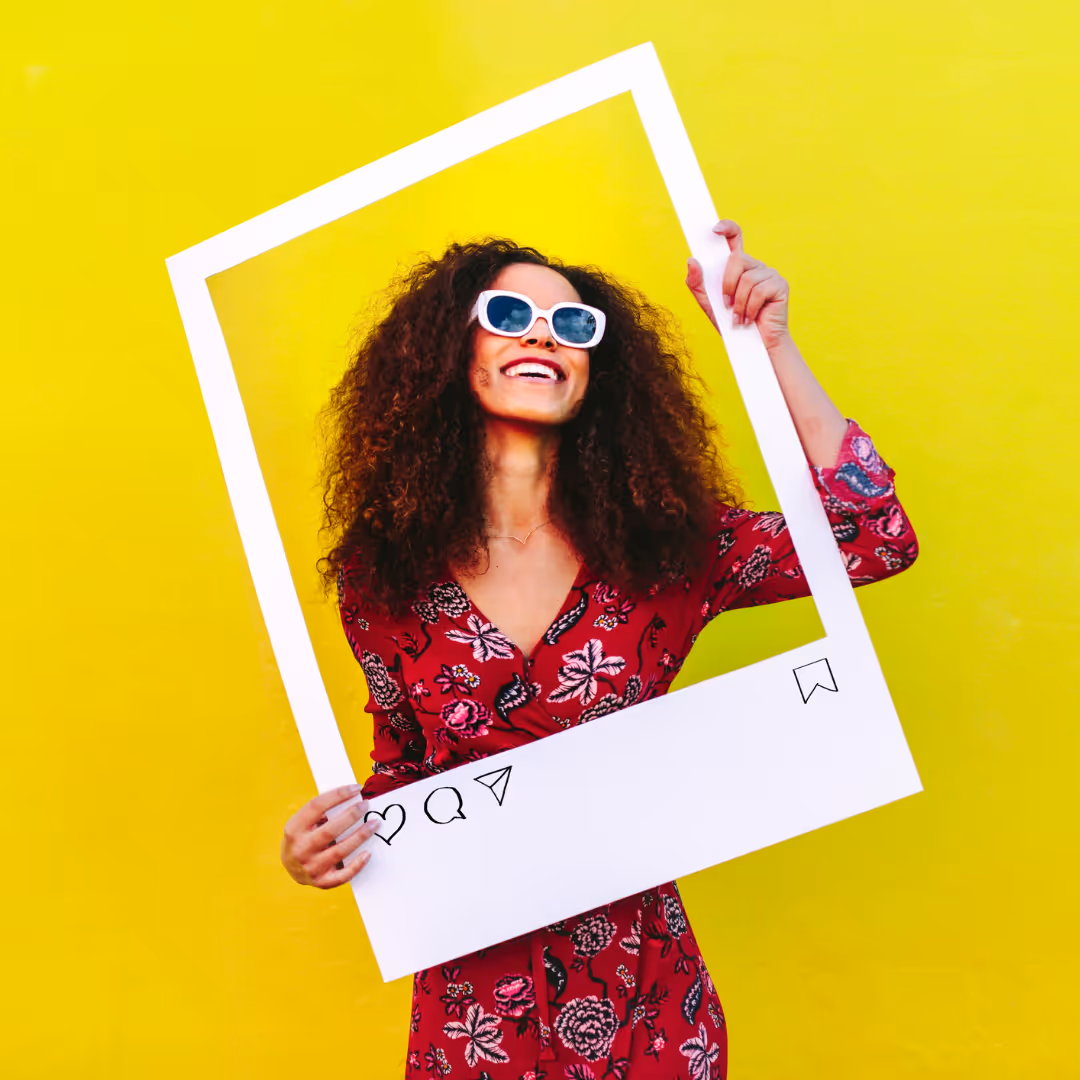Smiling woman with curly hair and white sunglasses holding a white Instagram-style photo frame against a yellow background.