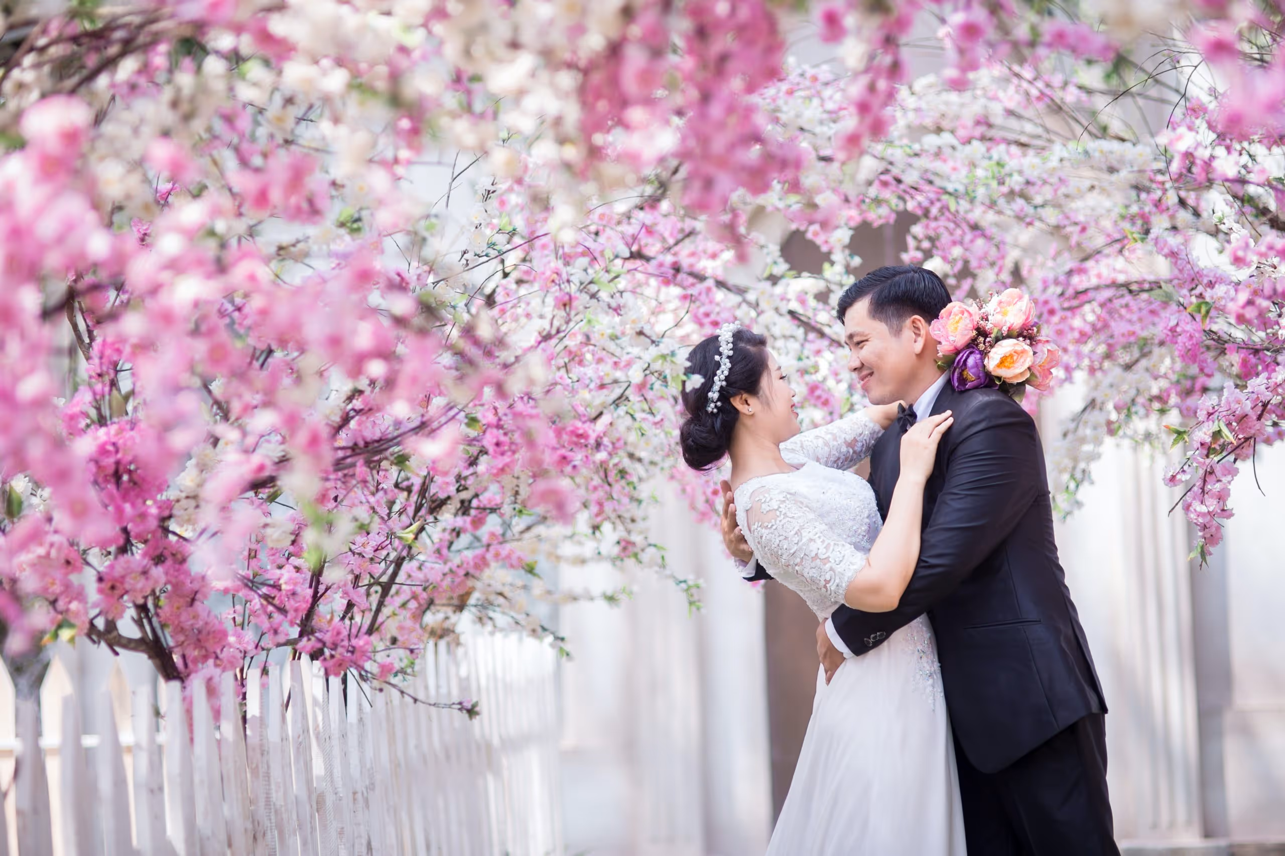 Bride and groom embracing and smiling under a canopy of pink and white cherry blossom flowers.