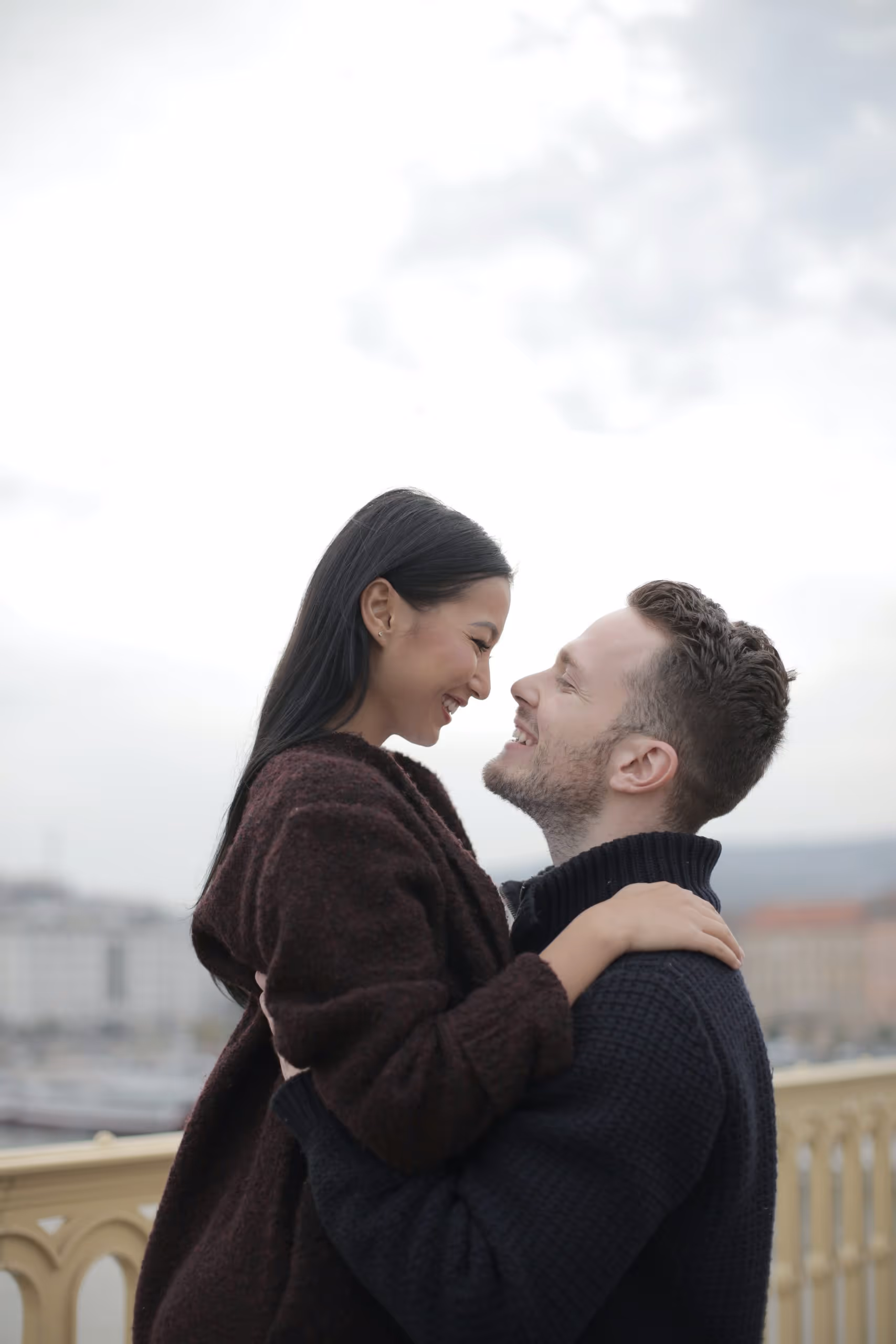 Couple smiling and embracing each other outdoors on a cloudy day.