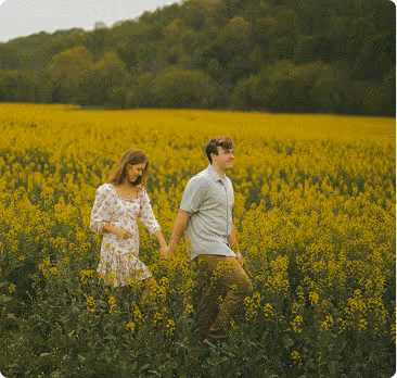 A couple holding hands walking through a yellow flower field with green trees in the background.