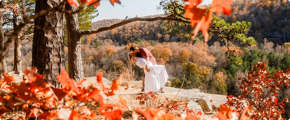 Couple kissing on a rocky ledge surrounded by autumn trees with orange and green foliage.