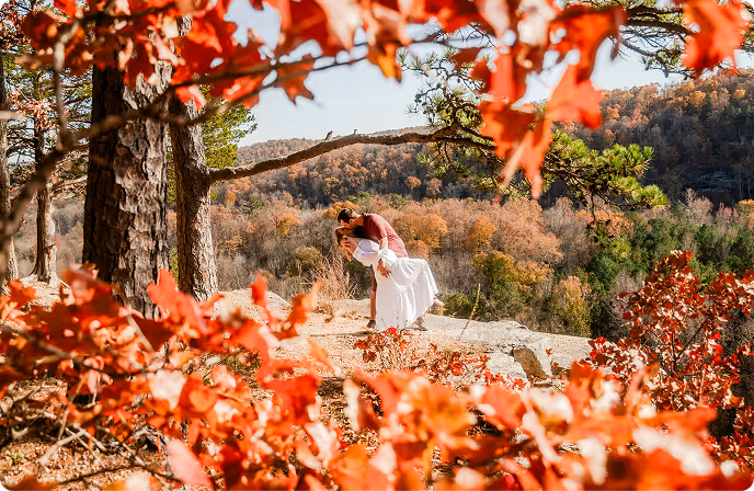 Couple kissing on a rock ledge surrounded by autumn trees with orange leaves and a forested mountain background.