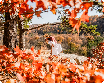 Couple in wedding attire embracing on a rock ledge surrounded by autumn foliage with a forested hill in the background.
