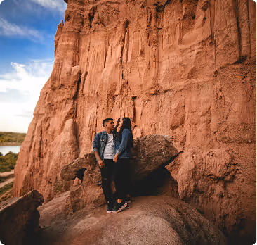 Couple standing close together on rocky terrain with tall red rock cliffs behind them under a partly cloudy sky.