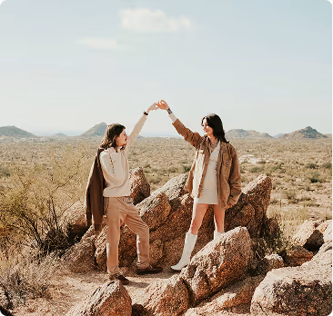 Two women on rocky terrain high-fiving with a vast desert landscape in the background.
