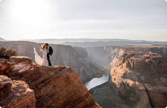 Couple in wedding attire standing on a rock ledge overlooking a canyon with a river below at sunset.