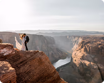 Bride and groom in wedding attire standing on a rocky cliff overlooking a canyon with a river below at sunset.
