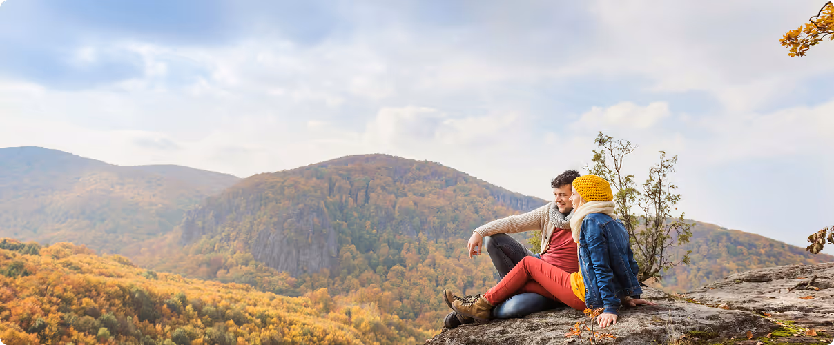 Couple sitting on a rocky ledge overlooking a forested mountain landscape in autumn.
