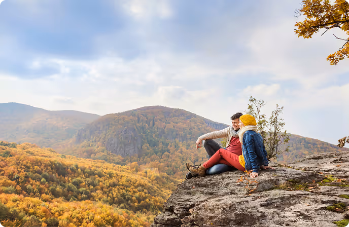 Couple sitting on a rocky cliff overlooking a forest with autumn foliage under a cloudy sky.