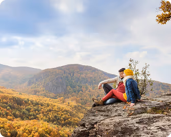 Couple sitting on a rocky cliff overlooking a forested mountain landscape in autumn.