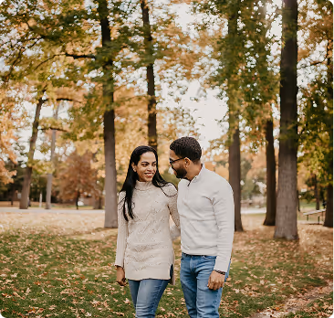 Smiling couple walking arm in arm in a park with autumn trees and fallen leaves.