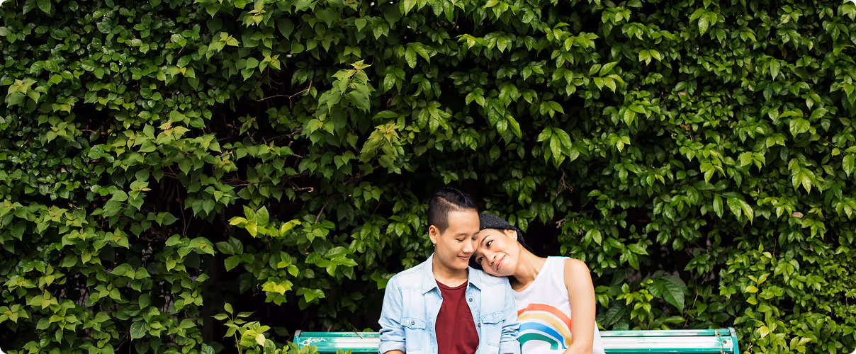 Couple sitting on a bench with a green leafy background, one resting their head on the other's shoulder.
