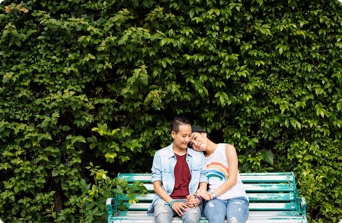 Couple sitting on a green bench, surrounded by lush green leaves, woman resting her head on man's shoulder.