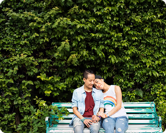 Couple sitting closely on a green bench in front of dense green foliage, holding hands and smiling.